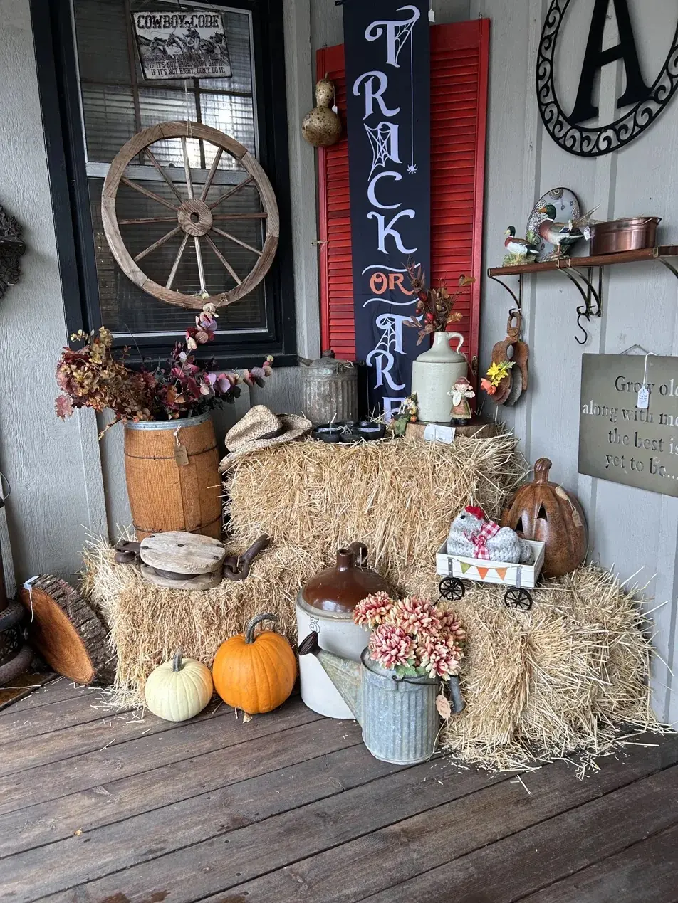 A fall display on a porch: hay bales with pumpkins, a wagon wheel, a stoneware crock, a galvanized watering can of mums, and a trick-or-treat sign